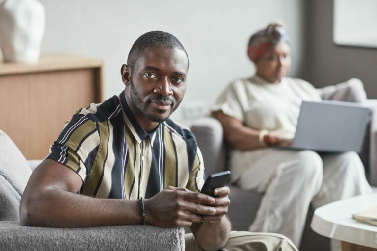 A confident African American man holding a smartphone while sitting indoors.