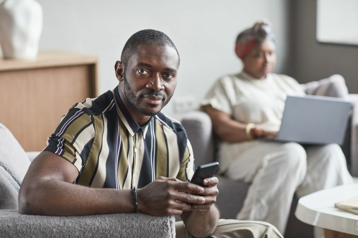 A confident African American man holding a smartphone while sitting indoors.